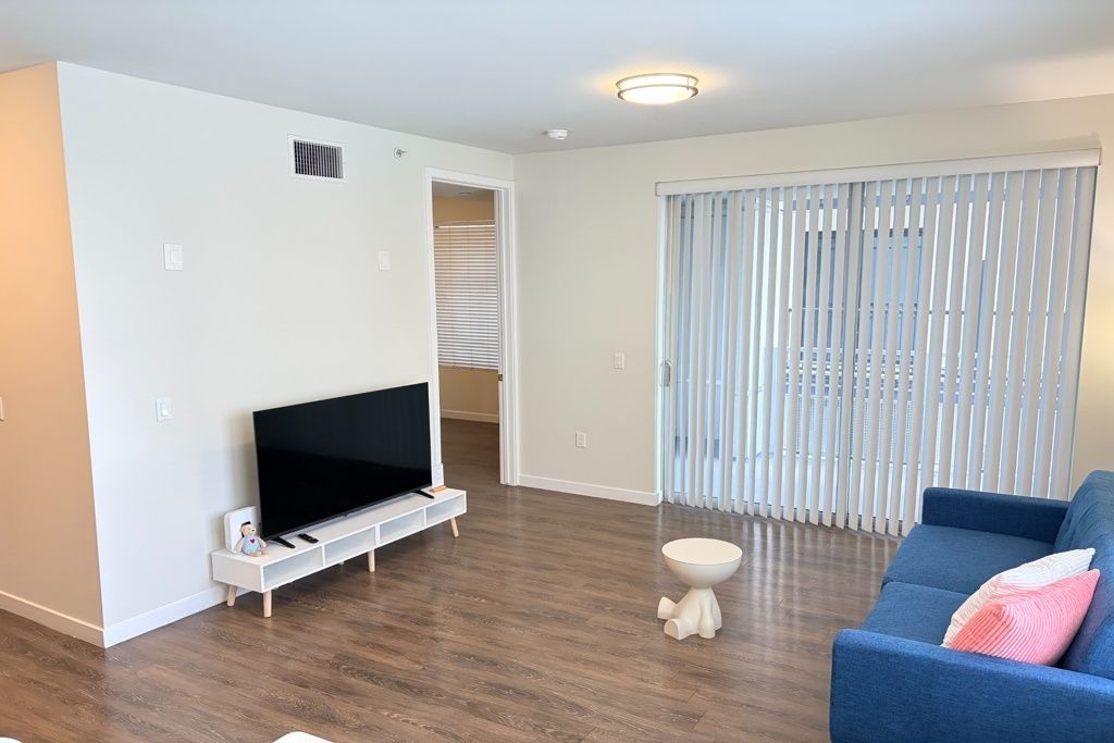 Bright living room with TV on stand, blue sofa, pink pillow, coffee table, and vertical blinds.