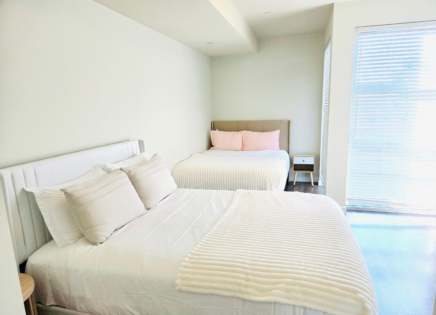 Bright bedroom with two white beds, beige headboard, and sunlight from a window blinds