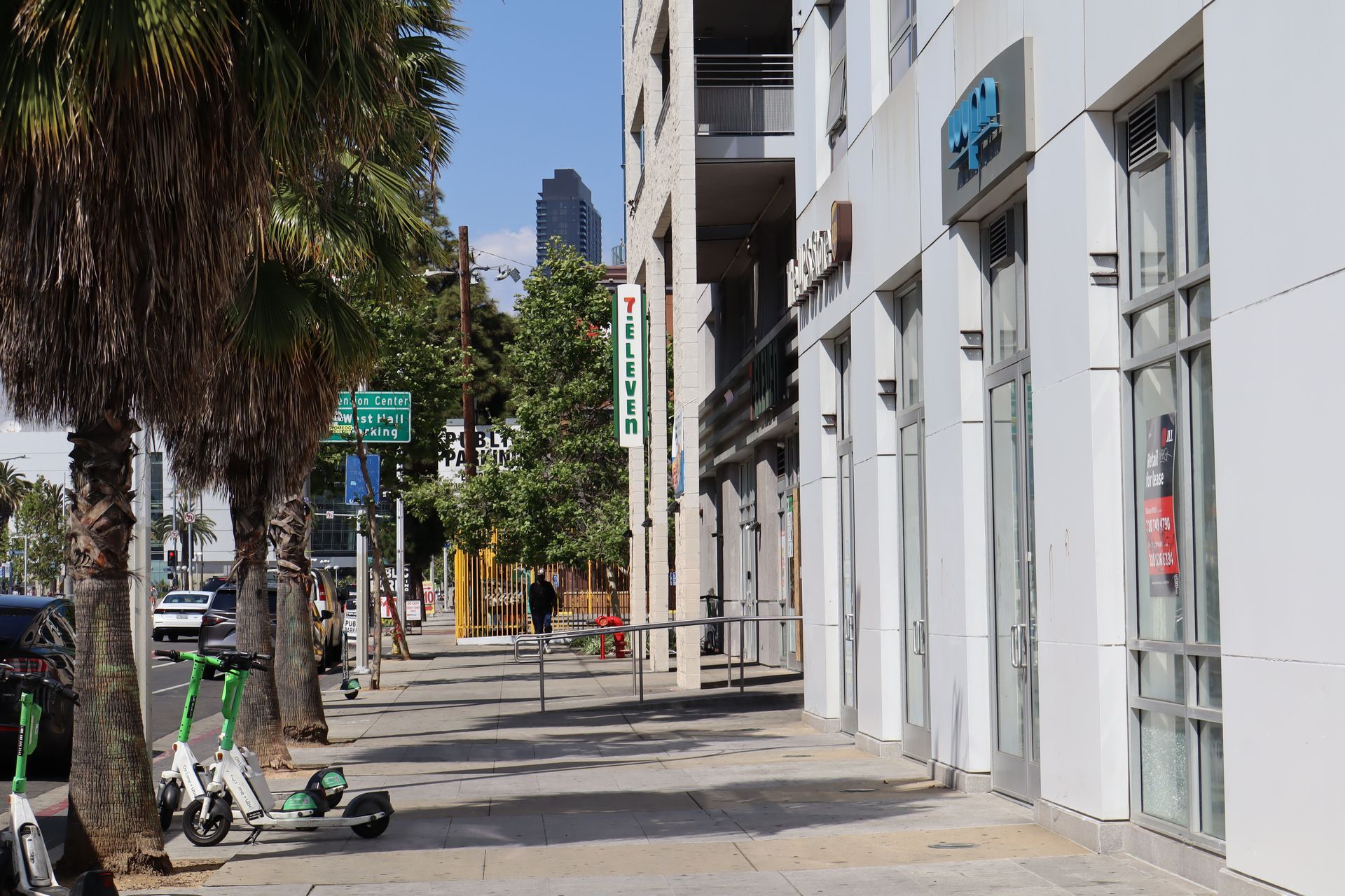Sunlit city sidewalk beside white buildings, with palm trees, parked scooters, and distant pedestrians.