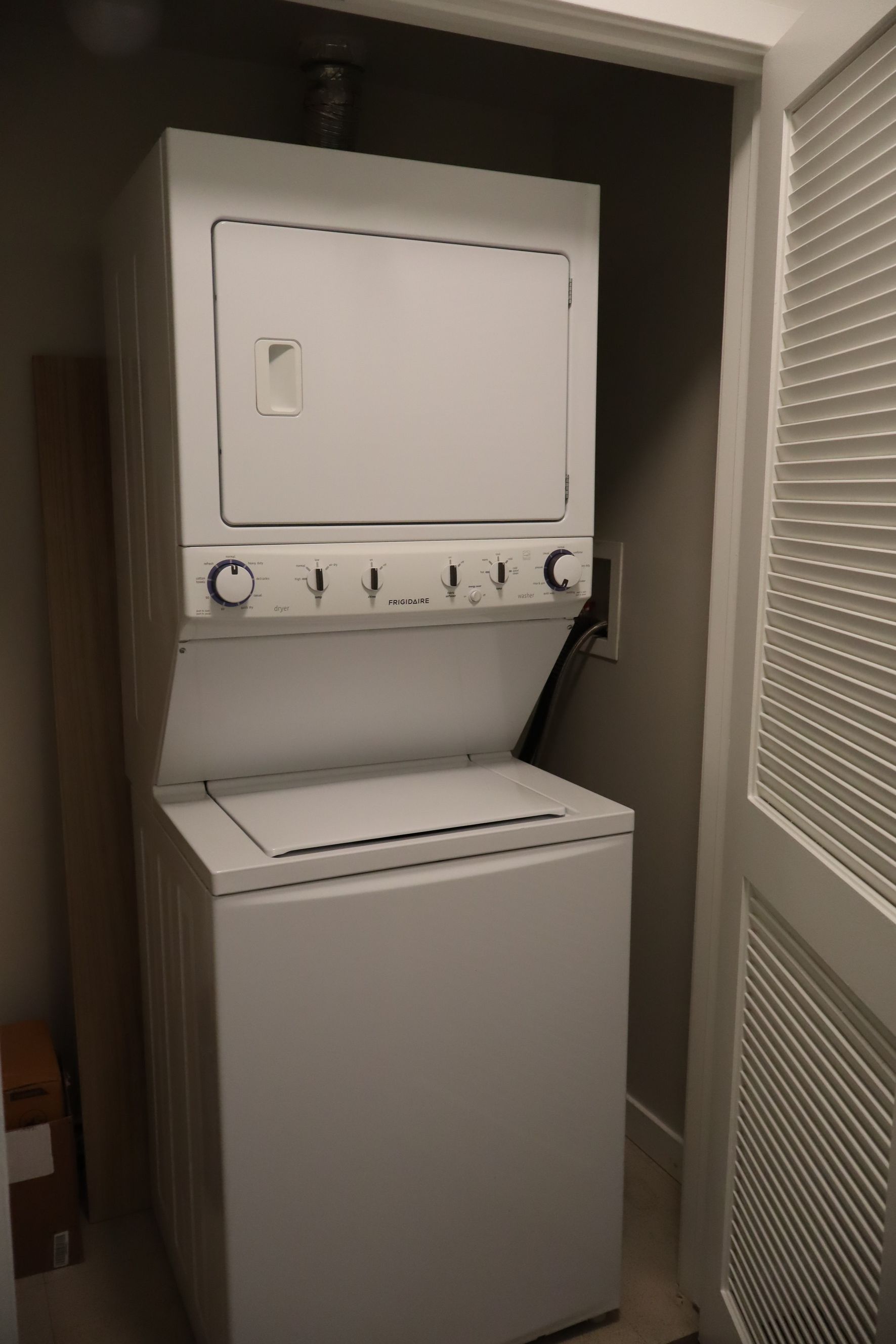 Stacked white washer and dryer in a narrow laundry closet with a window blind on the right.