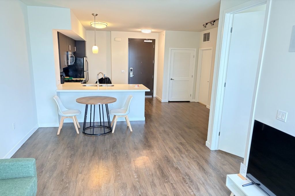Empty open-plan living-dining area with wood floors, white walls, and a small kitchen counter with stools.
