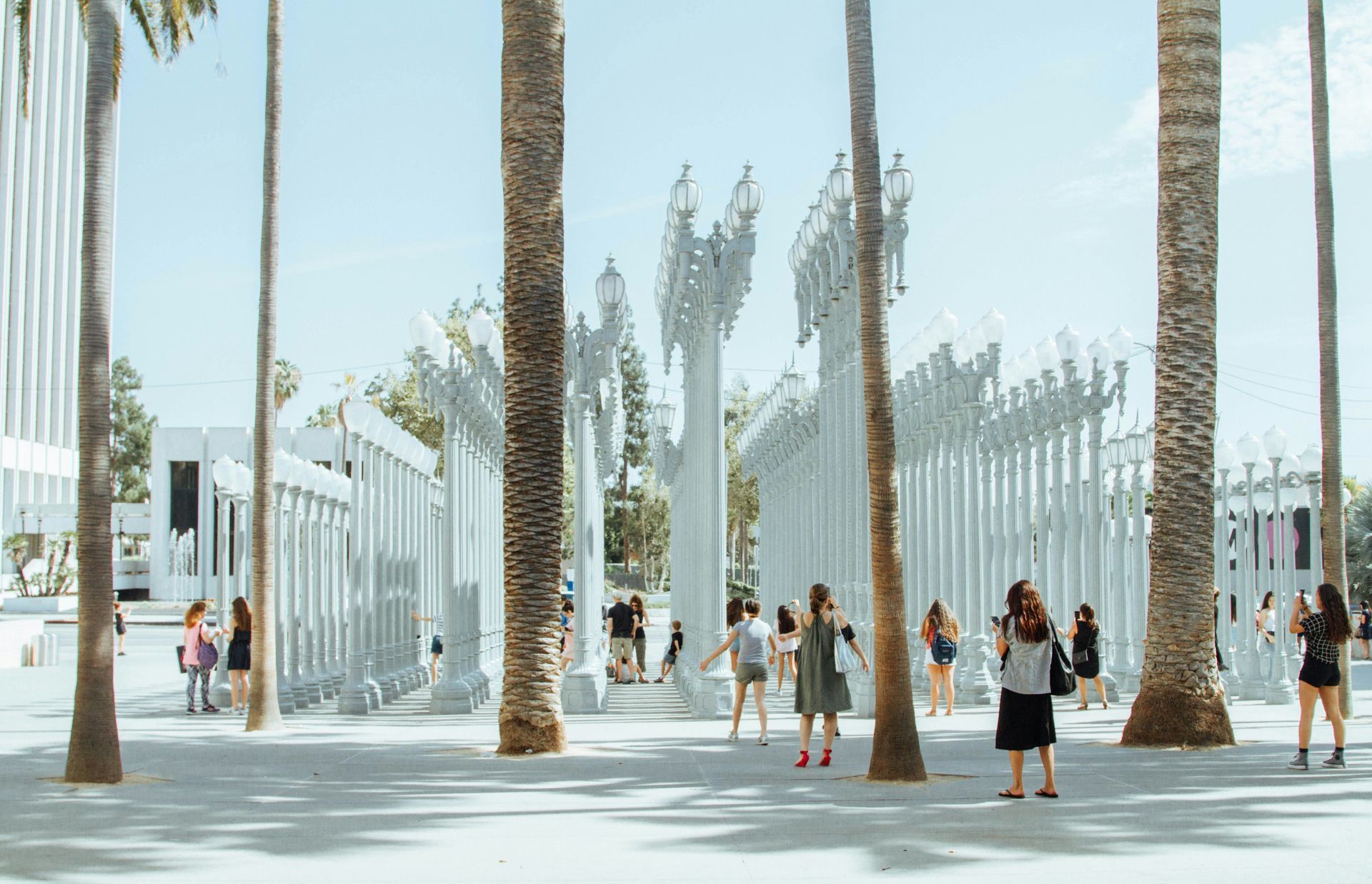 Sunny outdoor plaza with tall white sculptural columns and palm trees, with people walking and standing around