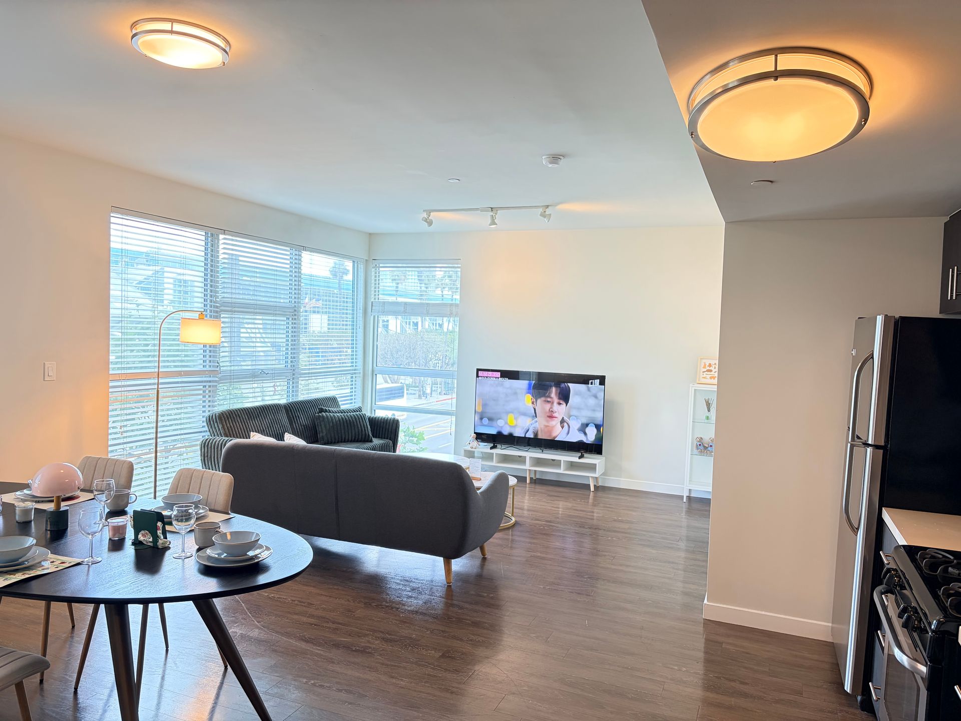 Bright living room with gray sofa, round table, TV, large windows, wood floor, and warm ceiling lights