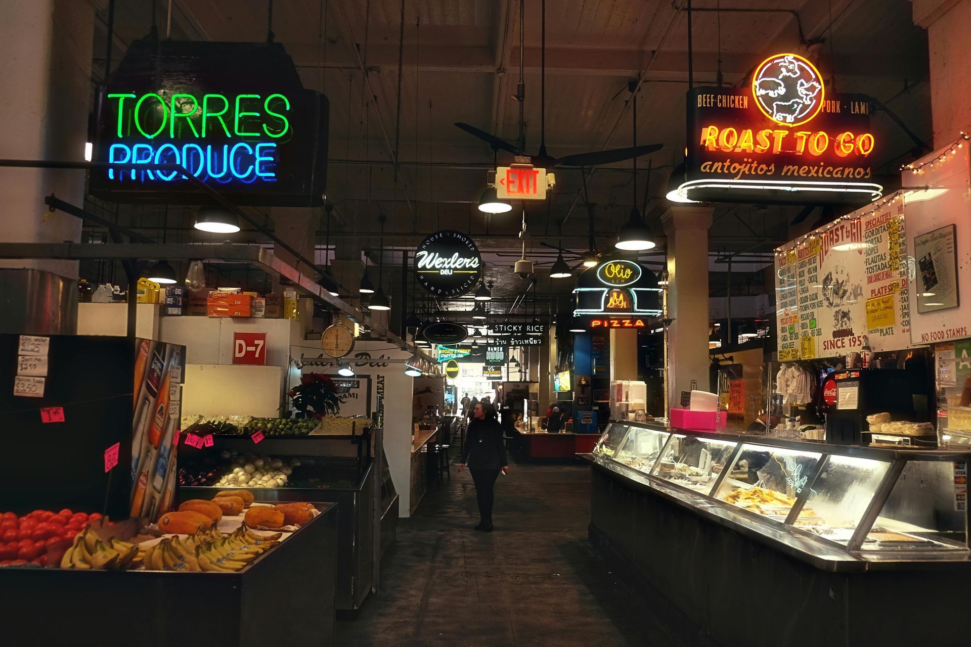 Indoor market aisle with produce stands, neon signs, and glass display cases under warm lighting