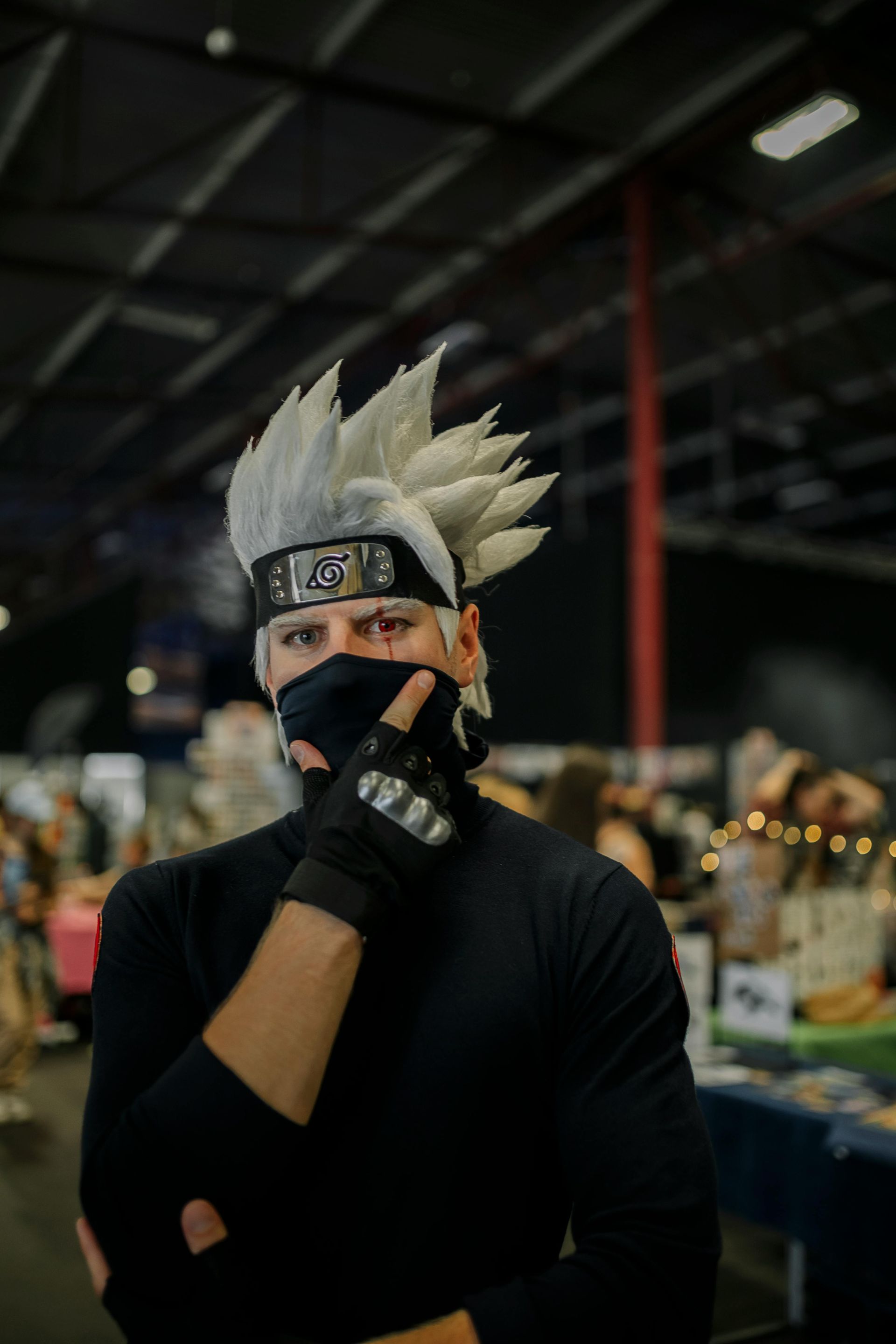 Masked cosplayer with spiky silver wig and black outfit at an indoor convention booth