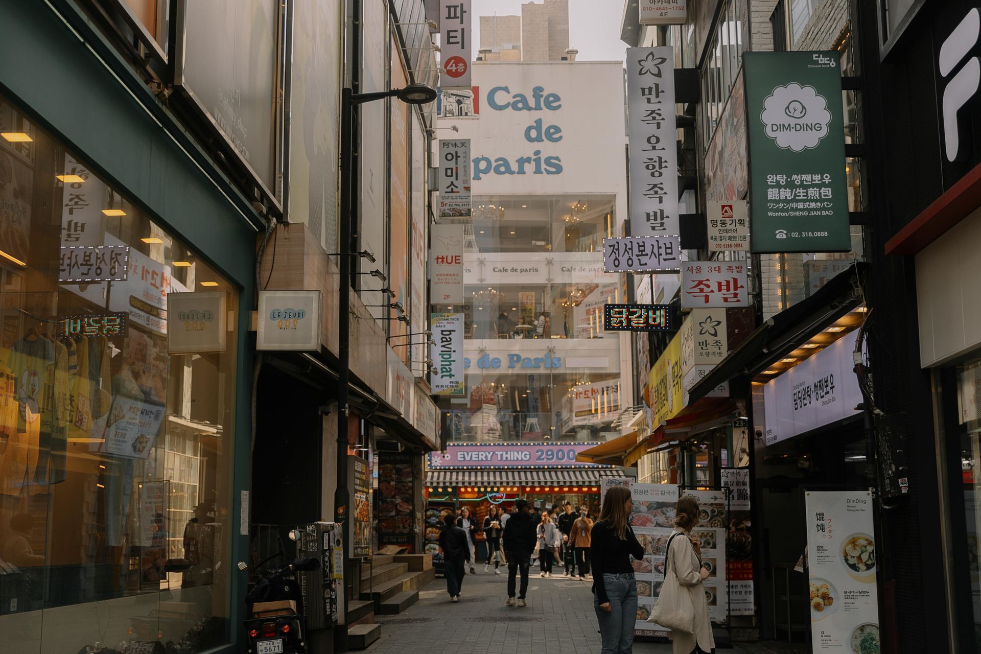 Narrow city alley with shops and hanging signs, including “Café de Paris,” and pedestrians walking below.
