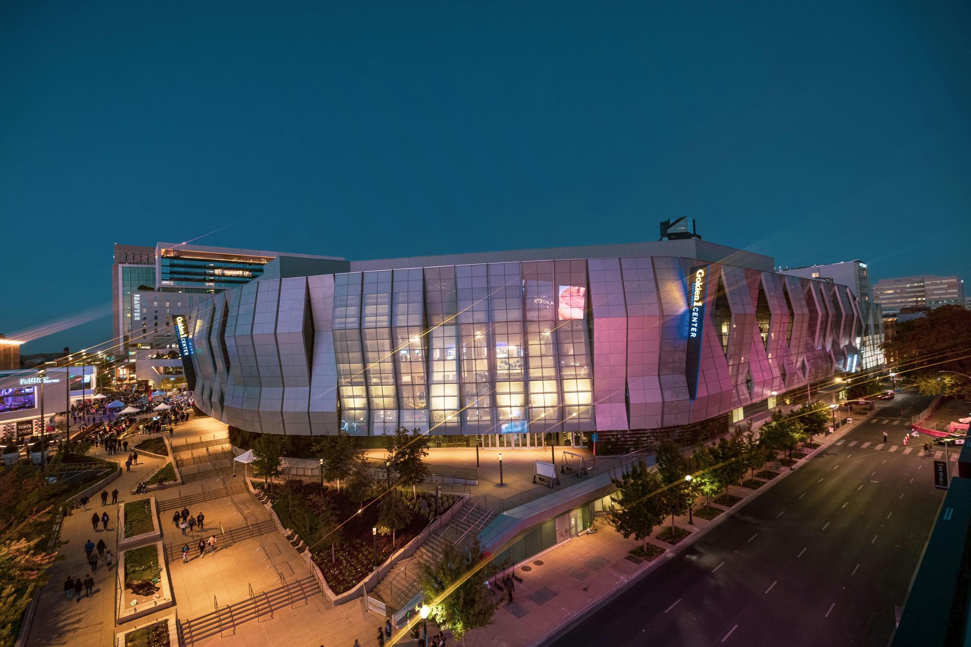 Modern glass building lit pink and blue at dusk, seen from above beside a road and trees