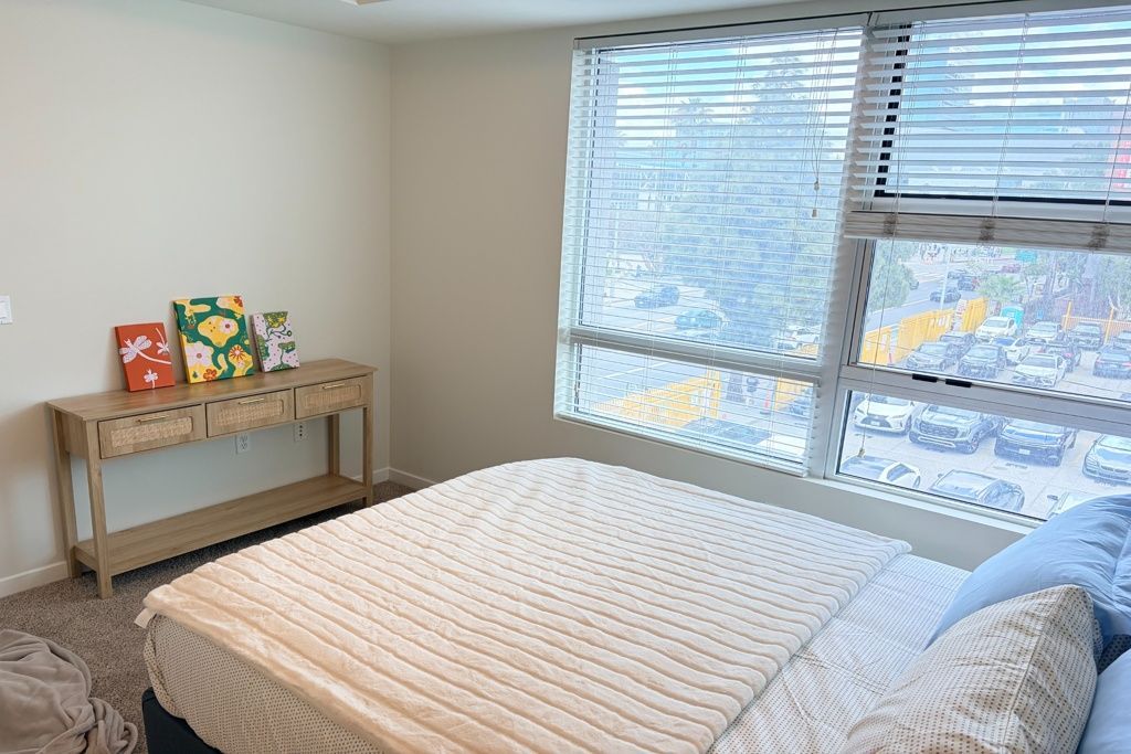 Bright bedroom with a bed, small wooden table, and large windows with blinds