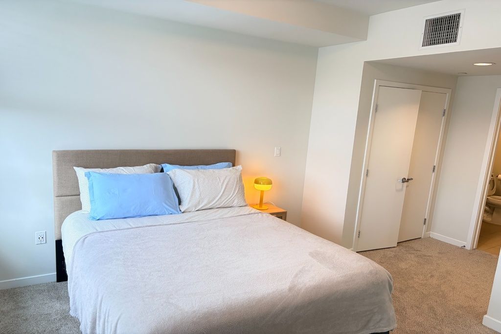 Bright bedroom with a bed, blue and white pillows, and a lit bedside lamp near an open closet.