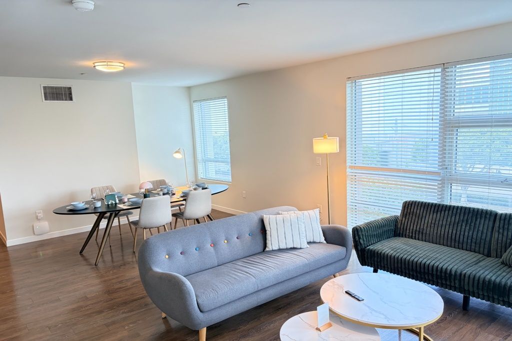 Bright modern living room with gray and dark striped sofas, round white table, dining area, and floor lamp.