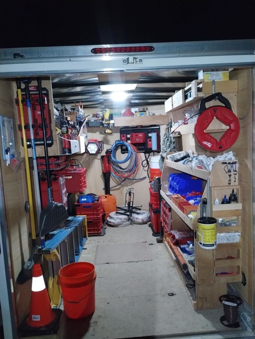 Narrow garage storage aisle with shelves, tools, red buckets, and coiled hoses under bright overhead light