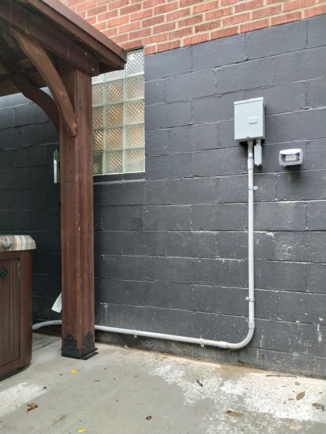 Gray cinderblock wall with electric meter, conduit, and a brown wooden post beside a concrete patio.