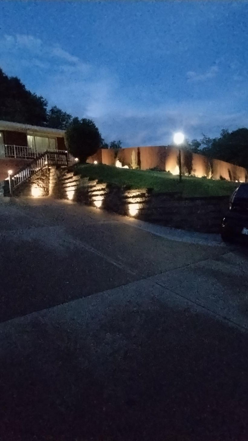 Dusk view of a lit walkway with steps, brick wall, trees, and blue evening sky