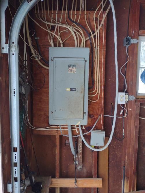Electrical breaker panel with exposed wiring in a wall cavity and a small white junction box below.