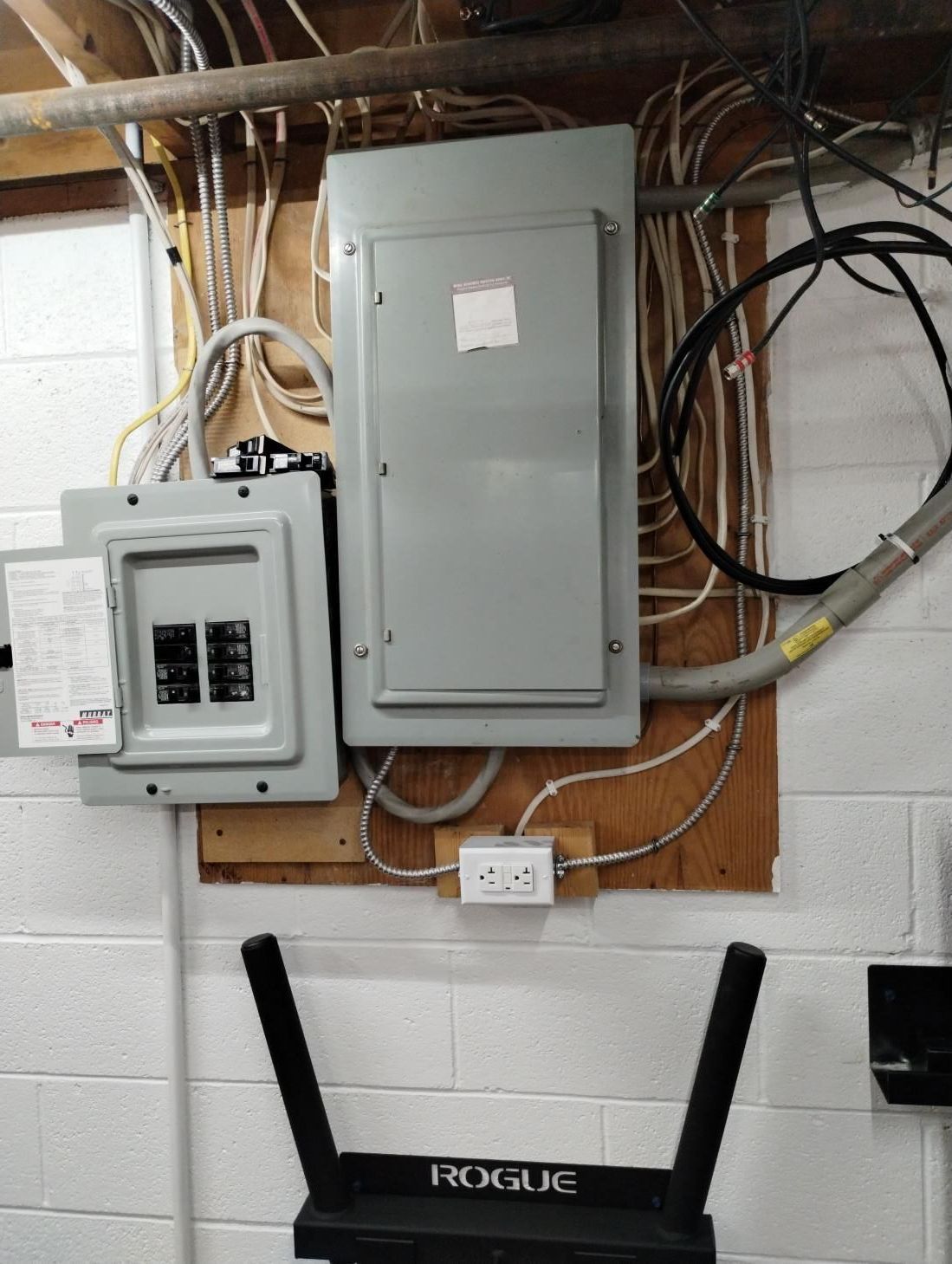Electrical panel and breaker box mounted on a cinderblock wall with exposed wiring and a white outlet below.