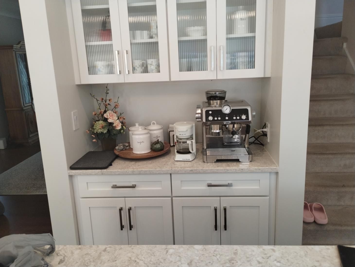 White kitchen coffee bar with glass cabinets, espresso machine, mugs, and floral decor