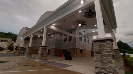 Covered outdoor patio with stone columns, white ceiling fans, and construction materials on a wet concrete floor