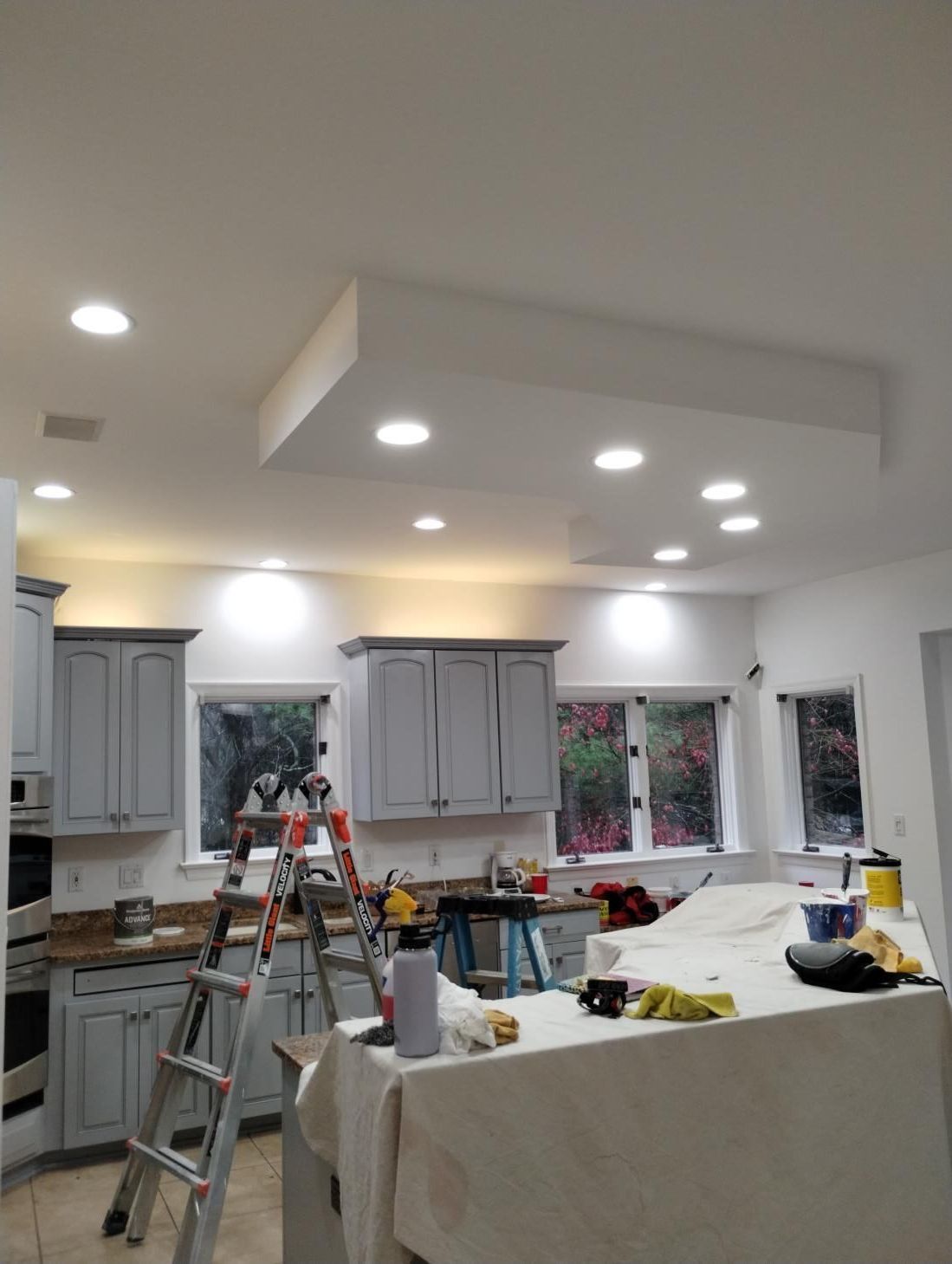 Kitchen under renovation with gray cabinets, island, ladders, tools, and recessed ceiling lights