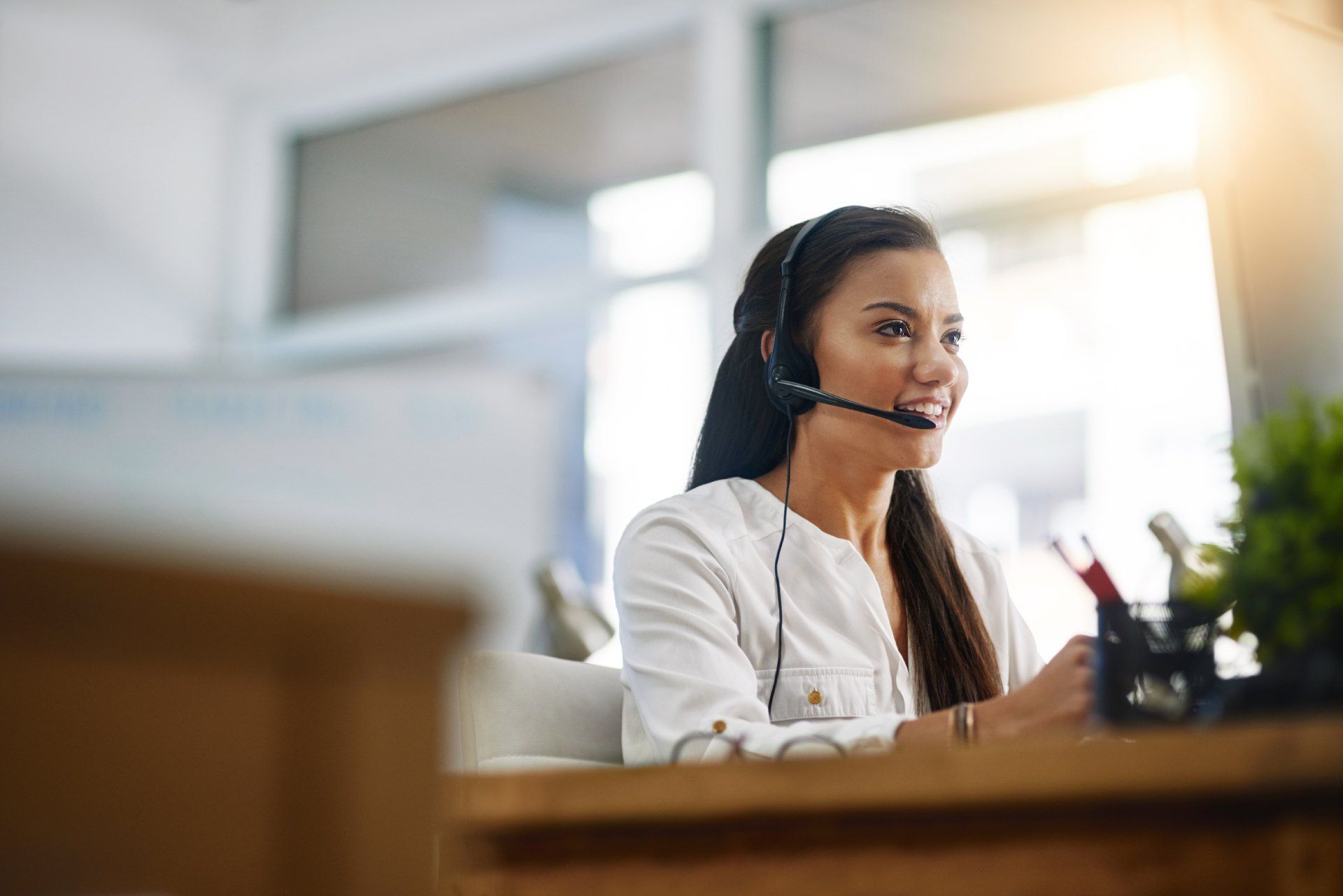A woman is wearing a headset and smiling while sitting at a desk.