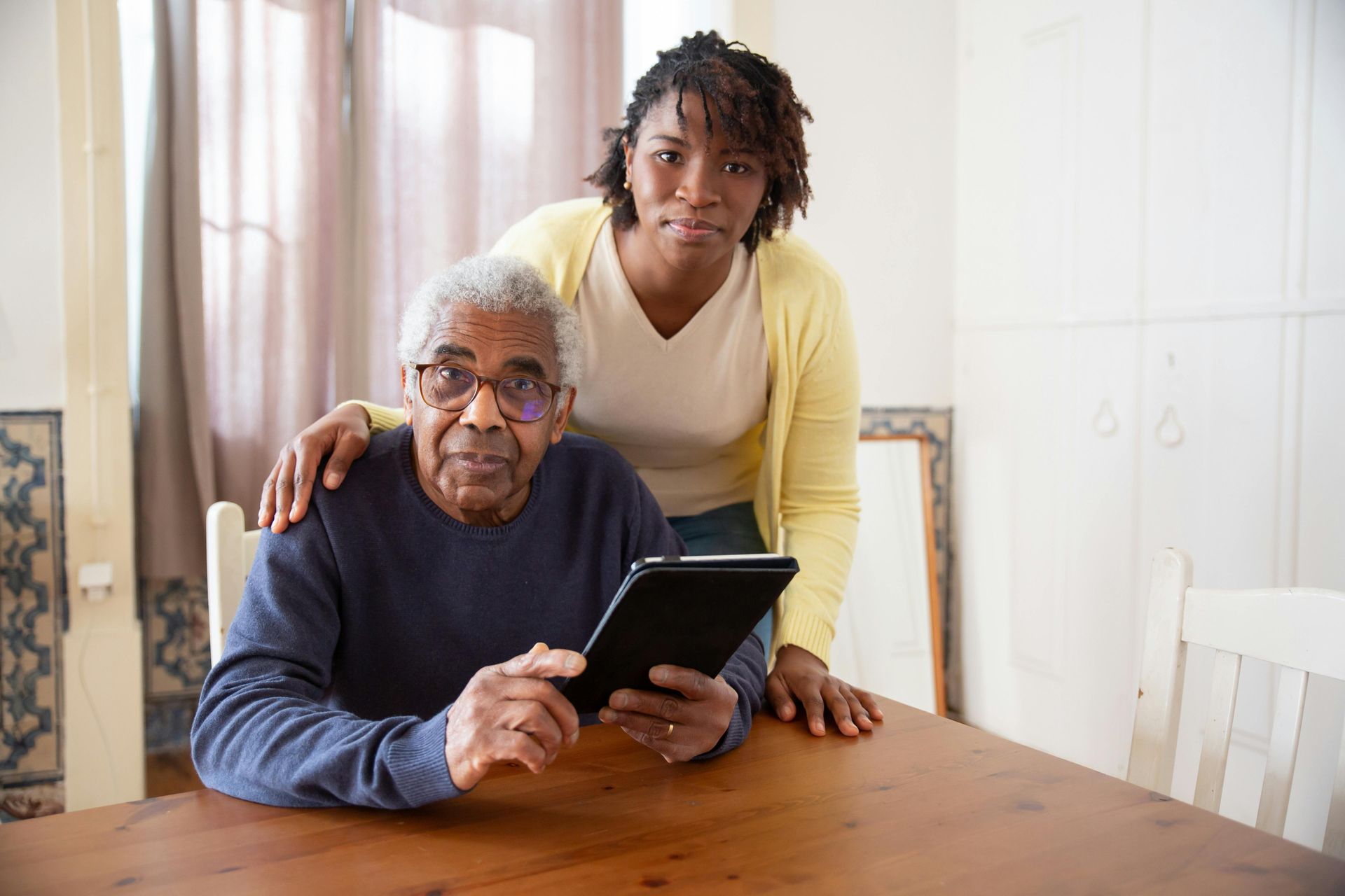 A person standing behind an older individual sitting at a wooden table while looking at a tablet together in a home.