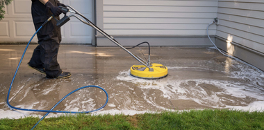 Person pressure washing a concrete driveway beside a house, with water spraying around a yellow surface cleaner.