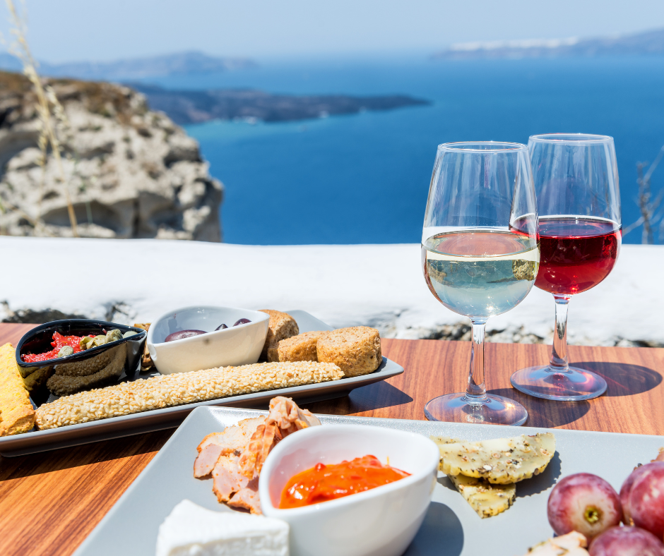 Two glasses of wine and a plate of food on a table with a view of the ocean.