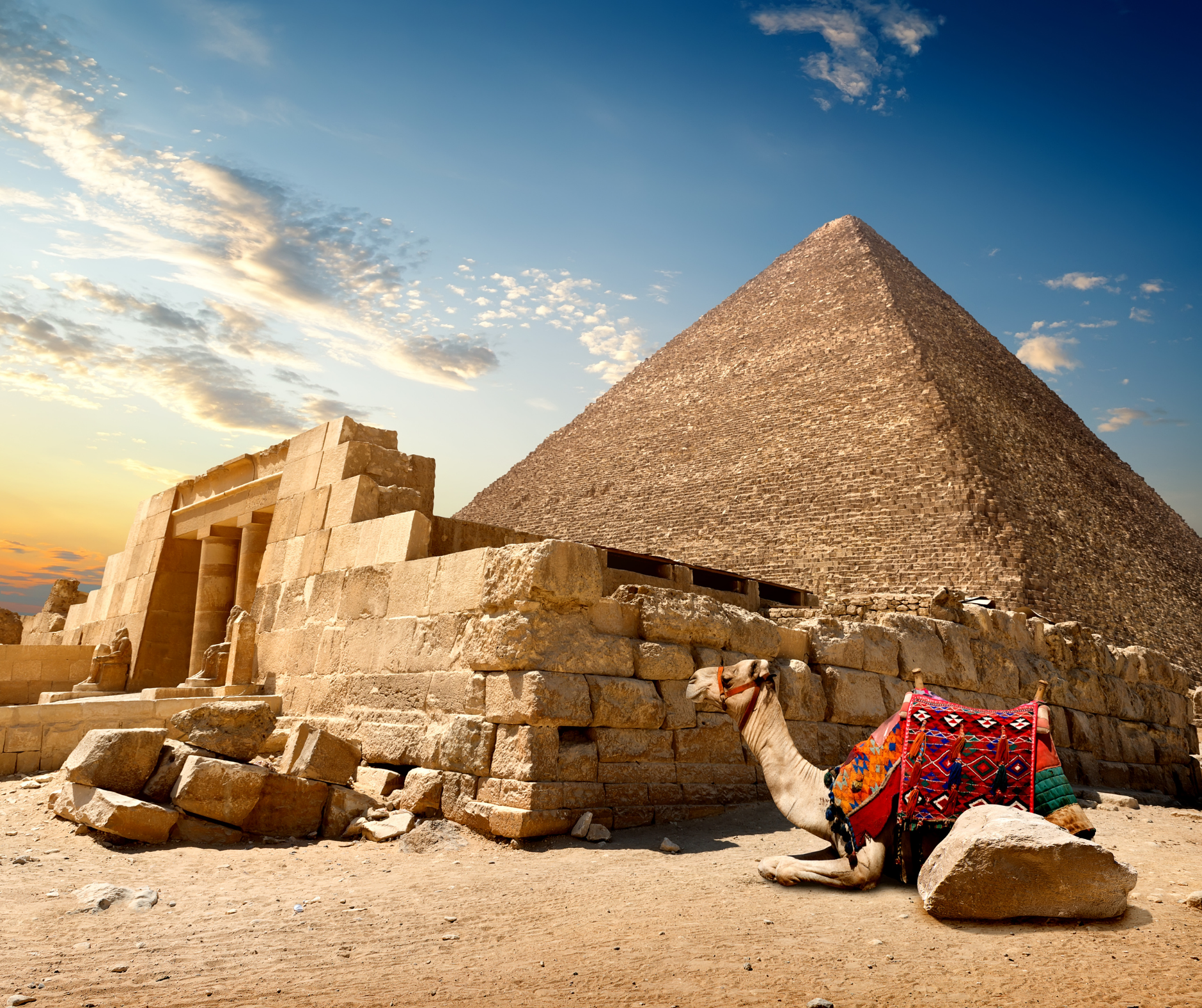 A camel is sitting in front of a pyramid in the desert.