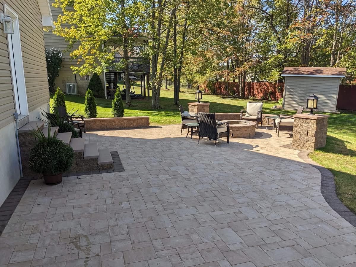 Backyard patio with fire pit, seating, and landscaping. Beige pavers, brown borders, and green grass. Sunny day.