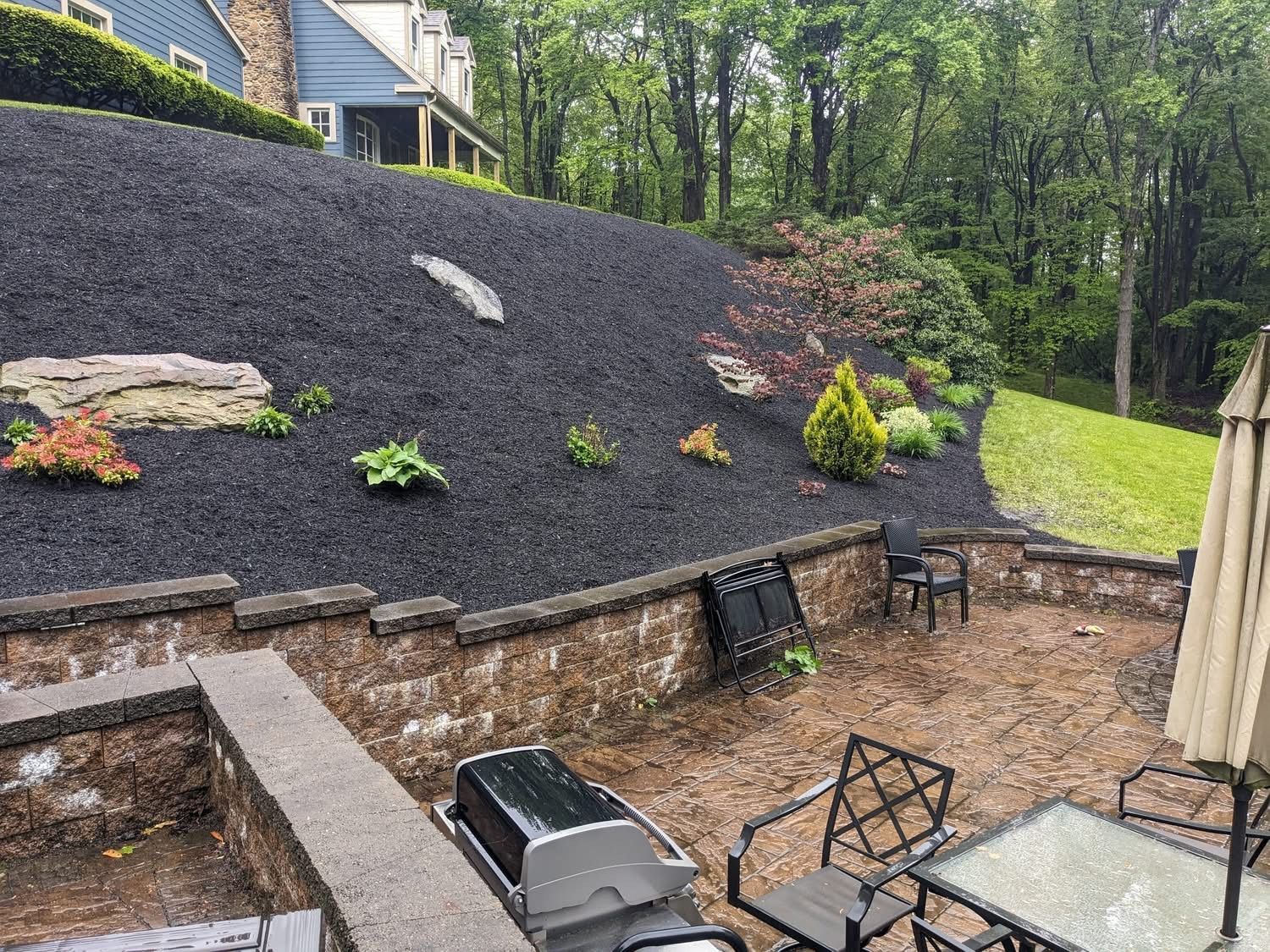 Patio with grill, table, chairs, and landscaped hillside covered in black mulch and plants.