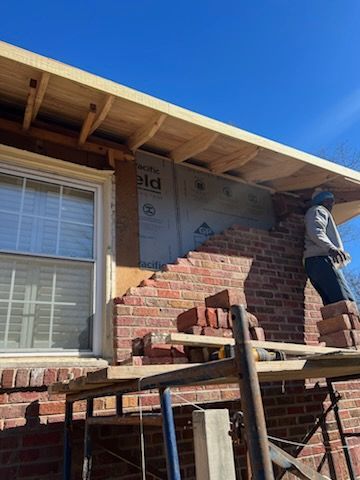 Bricklayer repairing a brick wall beneath a wooden eave on a sunny day.