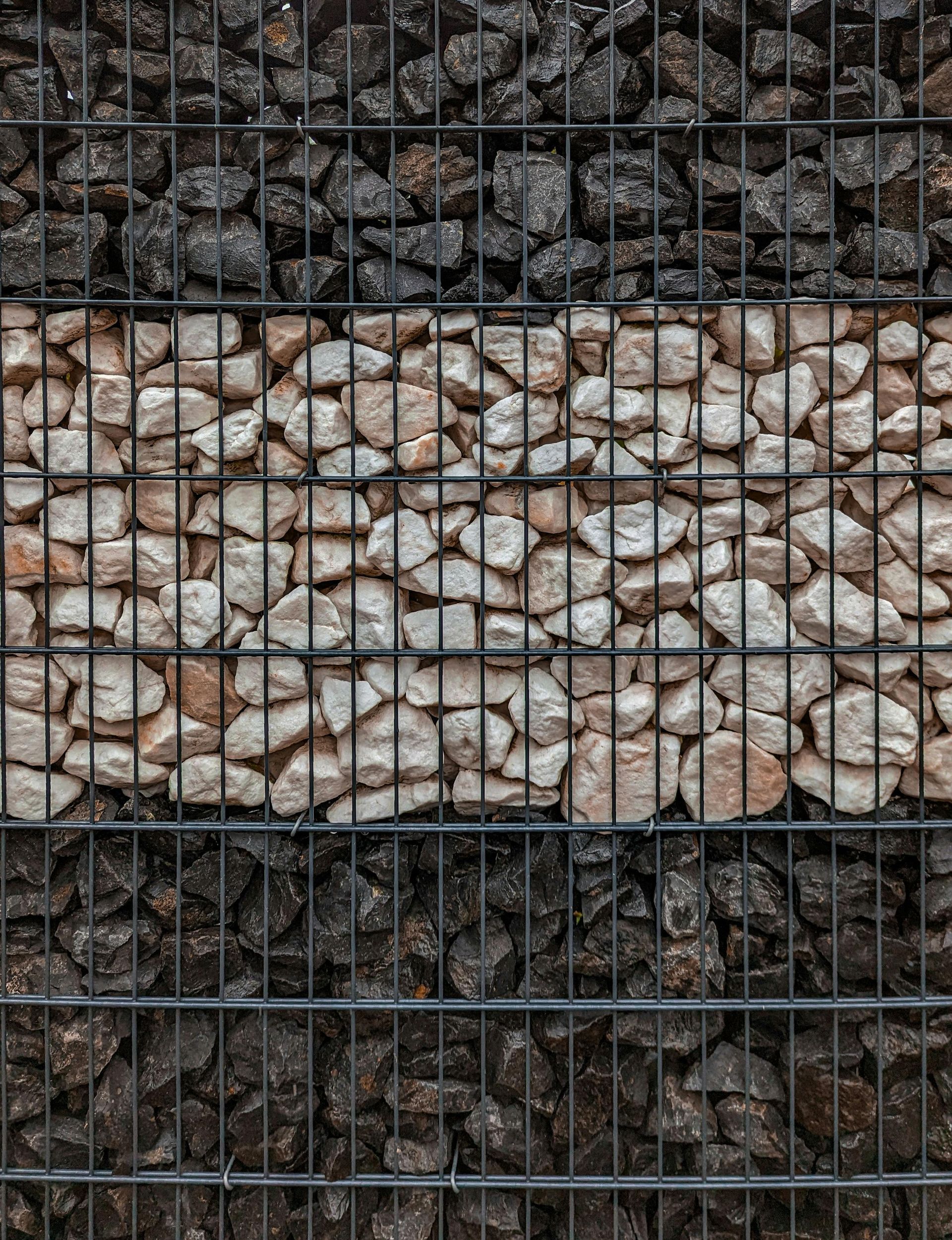 Sectioned gabion wall: black rocks at top and bottom, beige rocks in the middle, held in place by wire mesh.