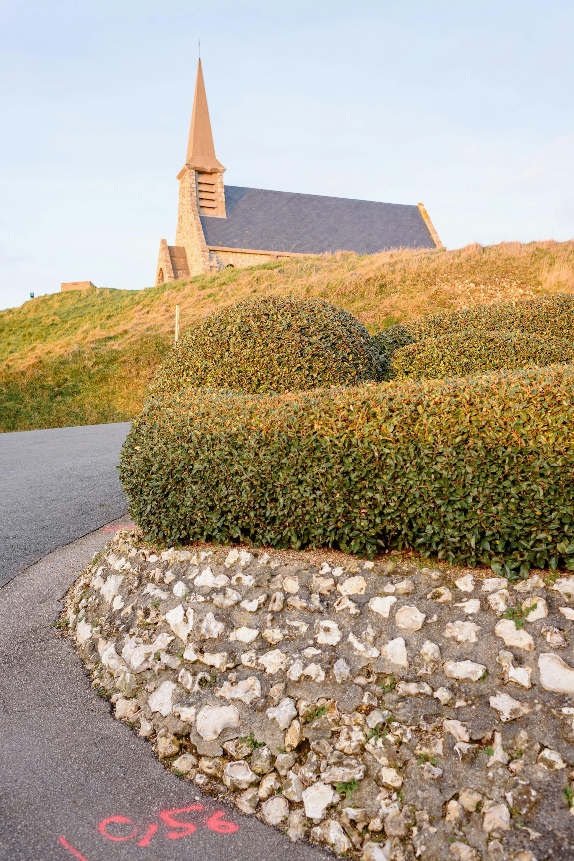 Church with steeple atop a grassy hill. Road and stone wall in foreground; blue sky.