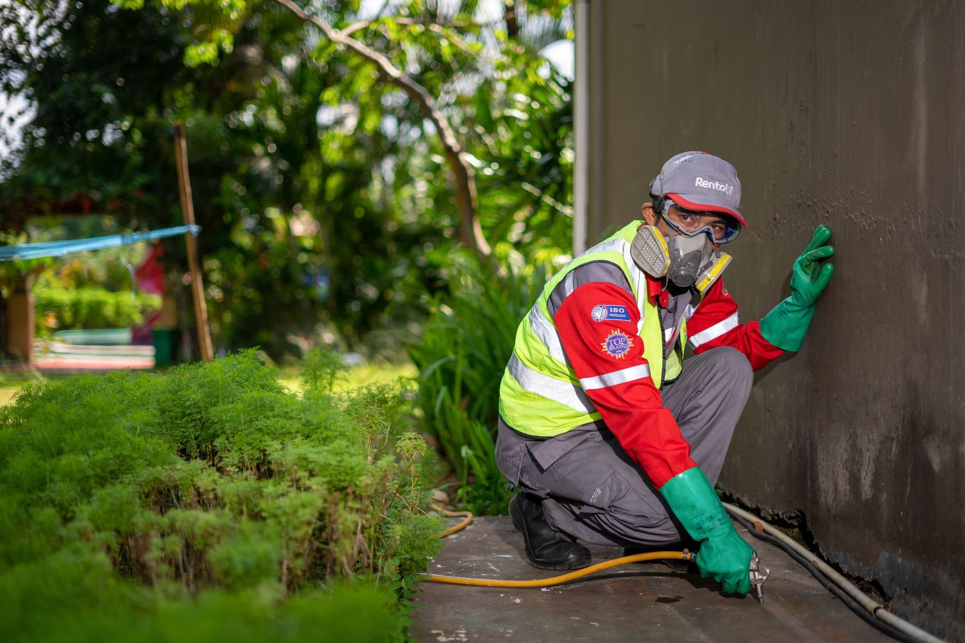 A worker in protective gear and a respirator crouches to spray a pesticide along a building foundation in a garden.