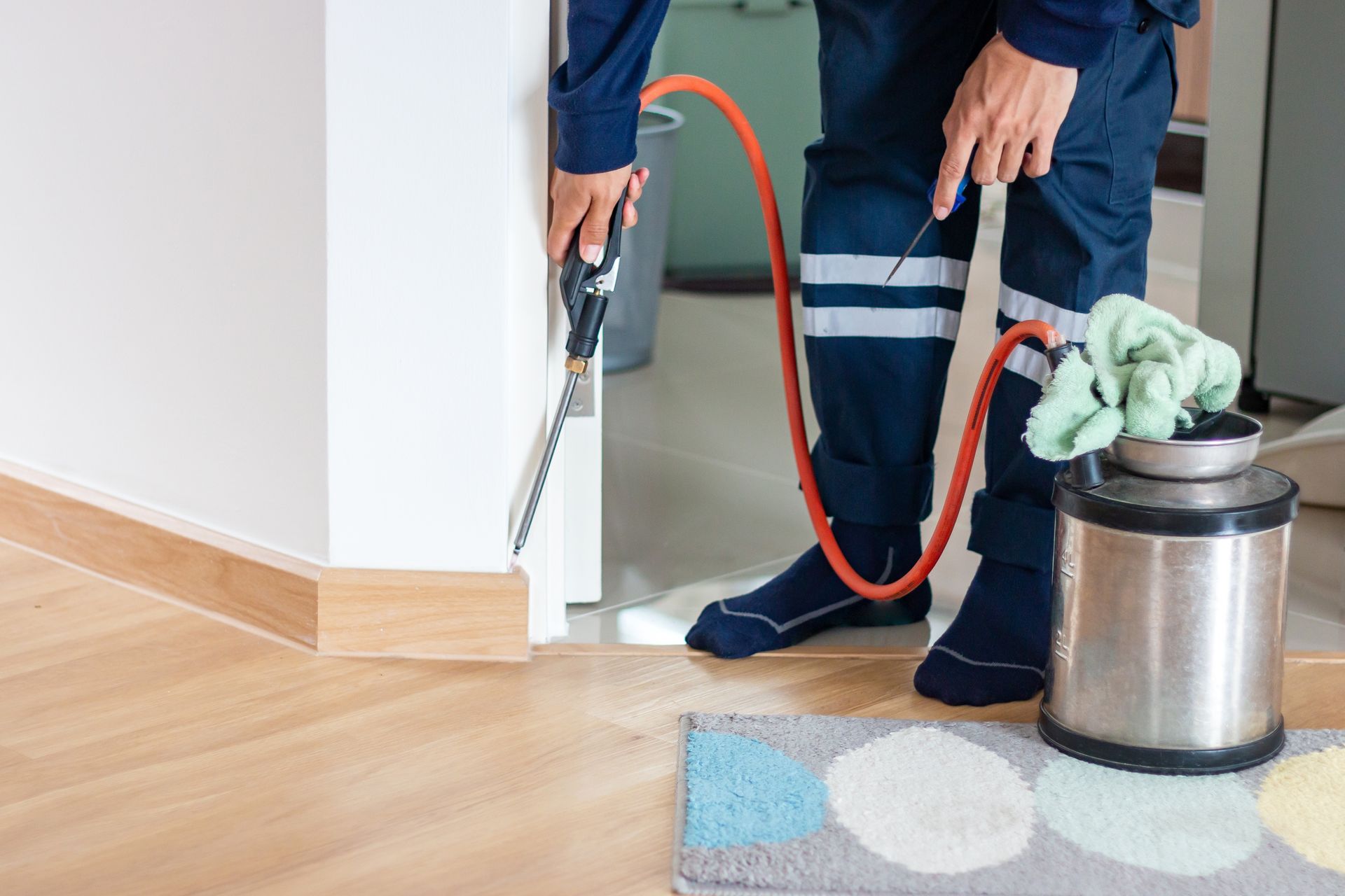 A professional wearing a uniform uses an orange hose and nozzle to spray pesticide along a doorway in a home.