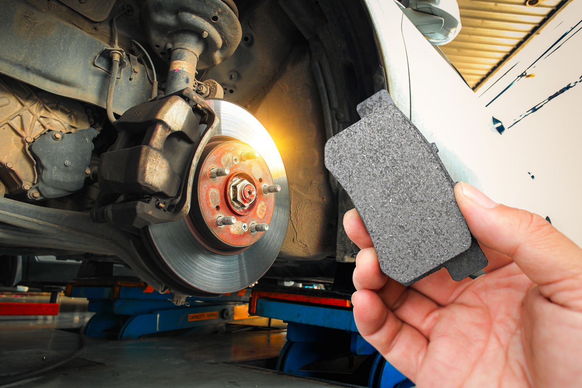 Hand holding a brake pad near a car wheel and exposed brake rotor in a repair shop