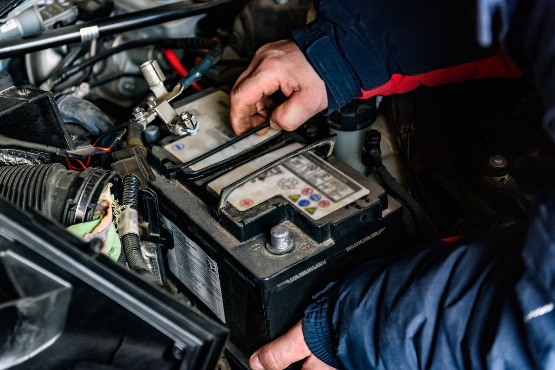 Mechanic working on a car battery under the hood, with tools and wiring visible.