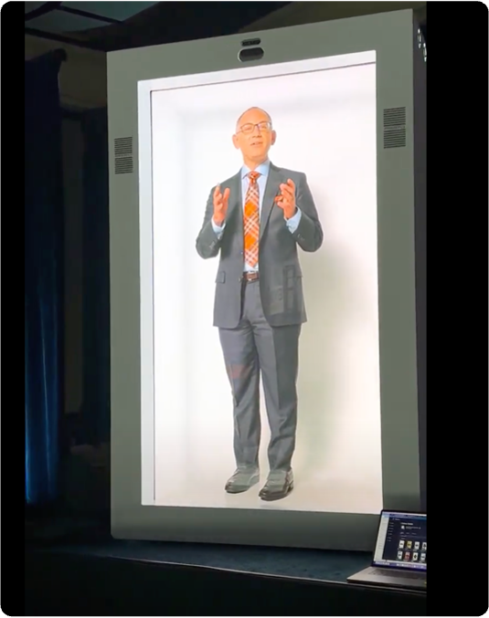A suited speaker stands on a large hologram display in a dark room, gesturing with both hands.