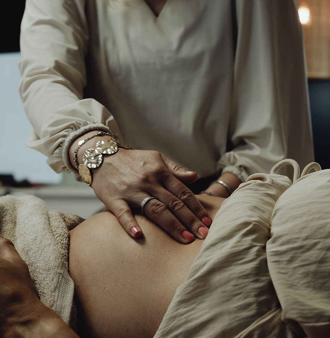 Handen masseren de buik van een persoon. Lichte huid, lichtgekleurde kleding, armbanden en een zichtbare ring. Beige handdoek en setting.