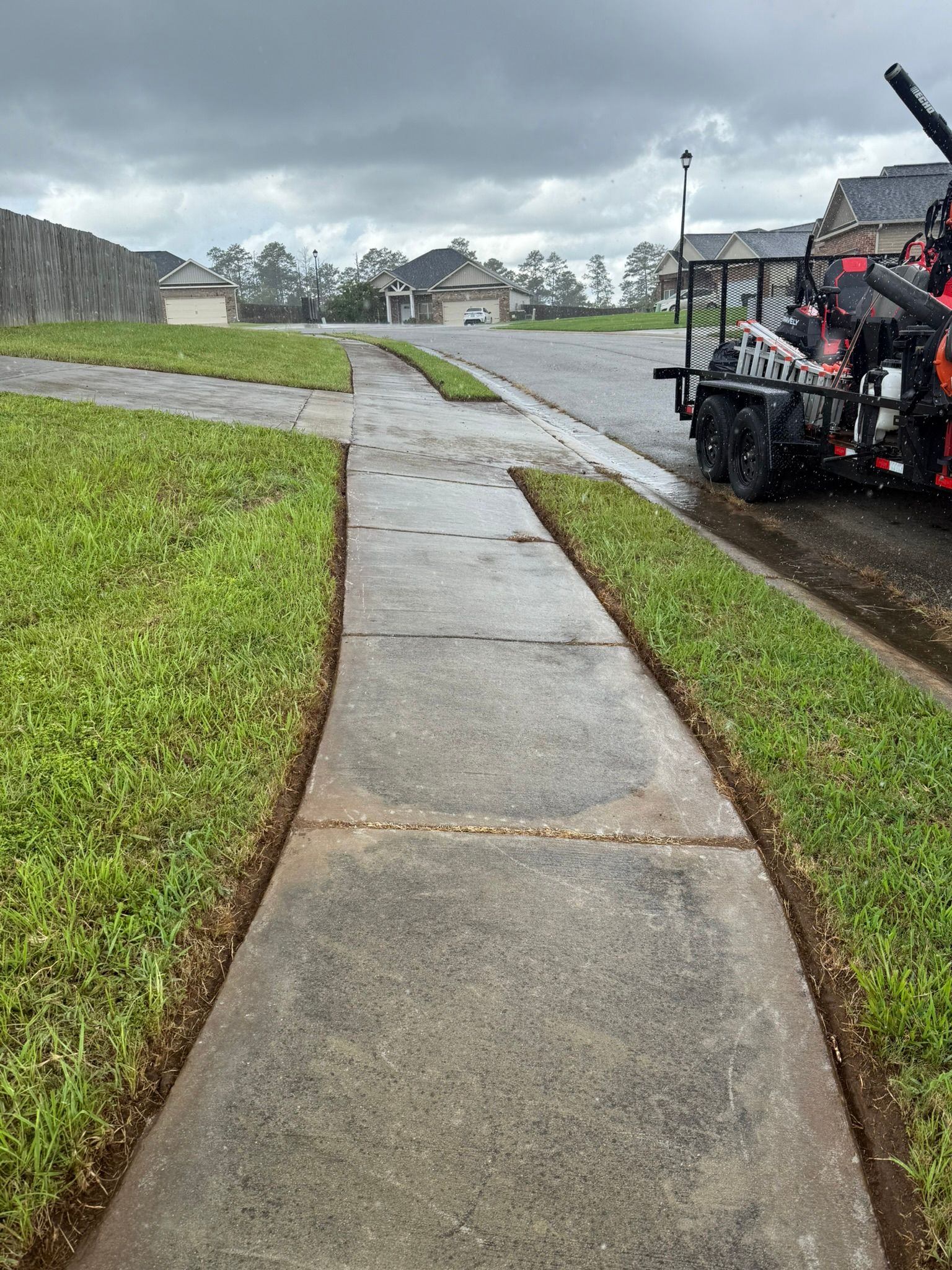Sidewalk bordered by green grass, leading towards houses on an overcast day.