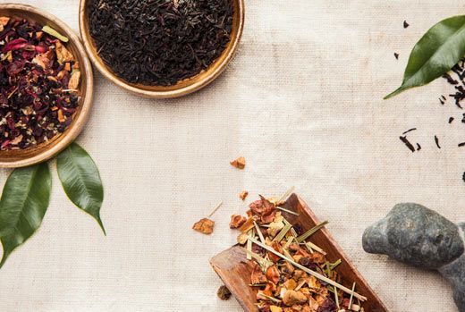 Bowls of tea leaves and herbs with green leaves on a beige linen surface.