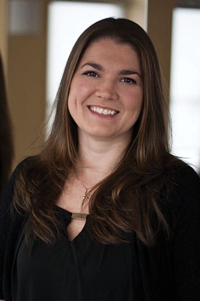 Woman with long brown hair smiles, wearing a black top and cardigan. Neutral background.