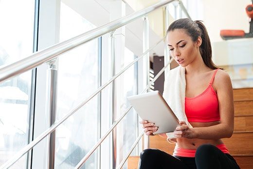 Woman in fitness attire, sits on stairs, looking at a tablet, towel on her shoulder.