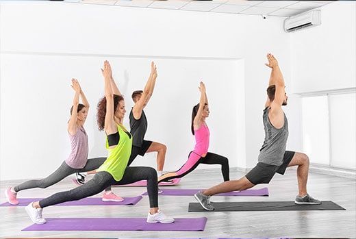 Group of people doing a yoga pose in a bright studio, arms raised, on purple mats.