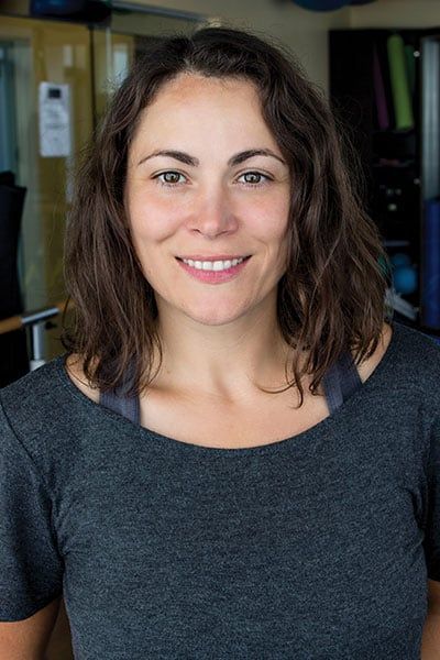 Woman smiling at the camera in a gym. Dark hair, grey shirt, neutral background.