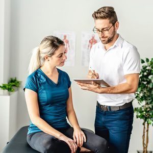 Physiotherapist with glasses examines a person sitting on an examination table, holding a tablet and pen.