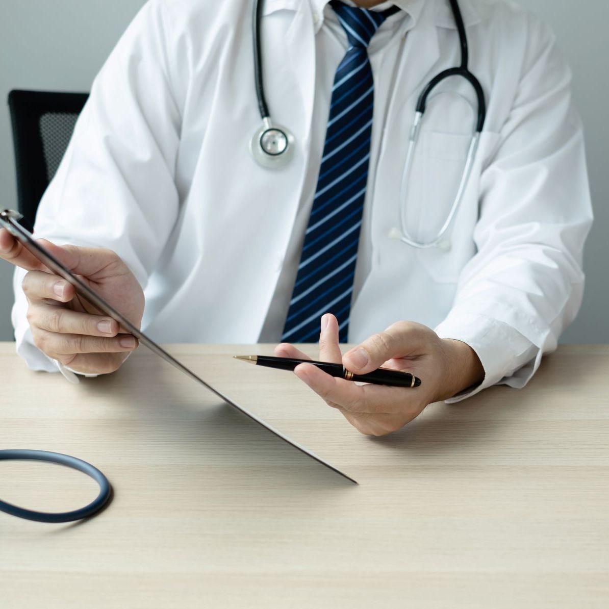 Doctor in white coat with stethoscope, holding a clipboard and pen at a desk.
