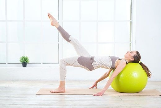 Woman doing a bridge pose on a mat, with one leg raised, resting head on a green exercise ball, near a window.