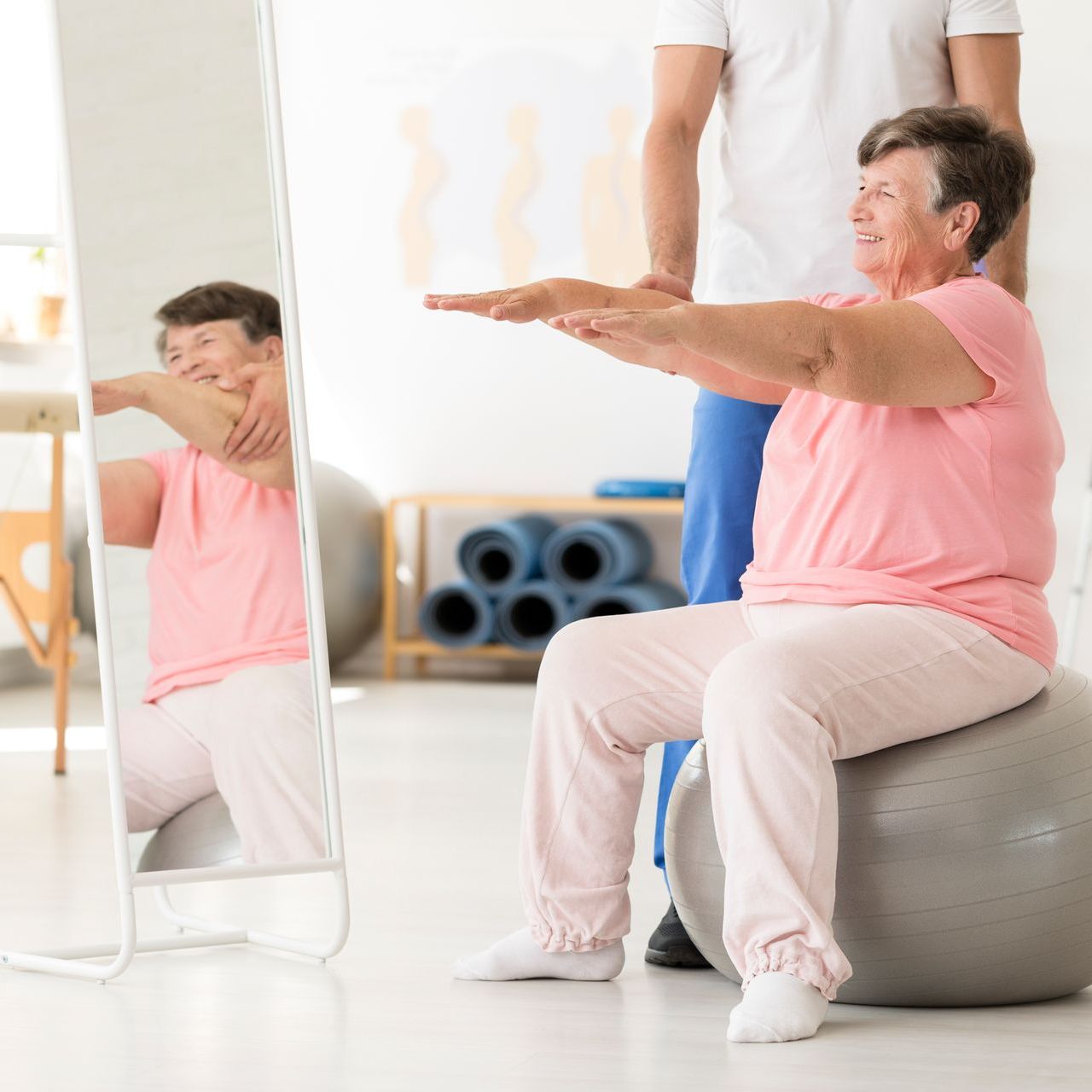 Woman seated on exercise ball stretches arms, guided by therapist in gym.
