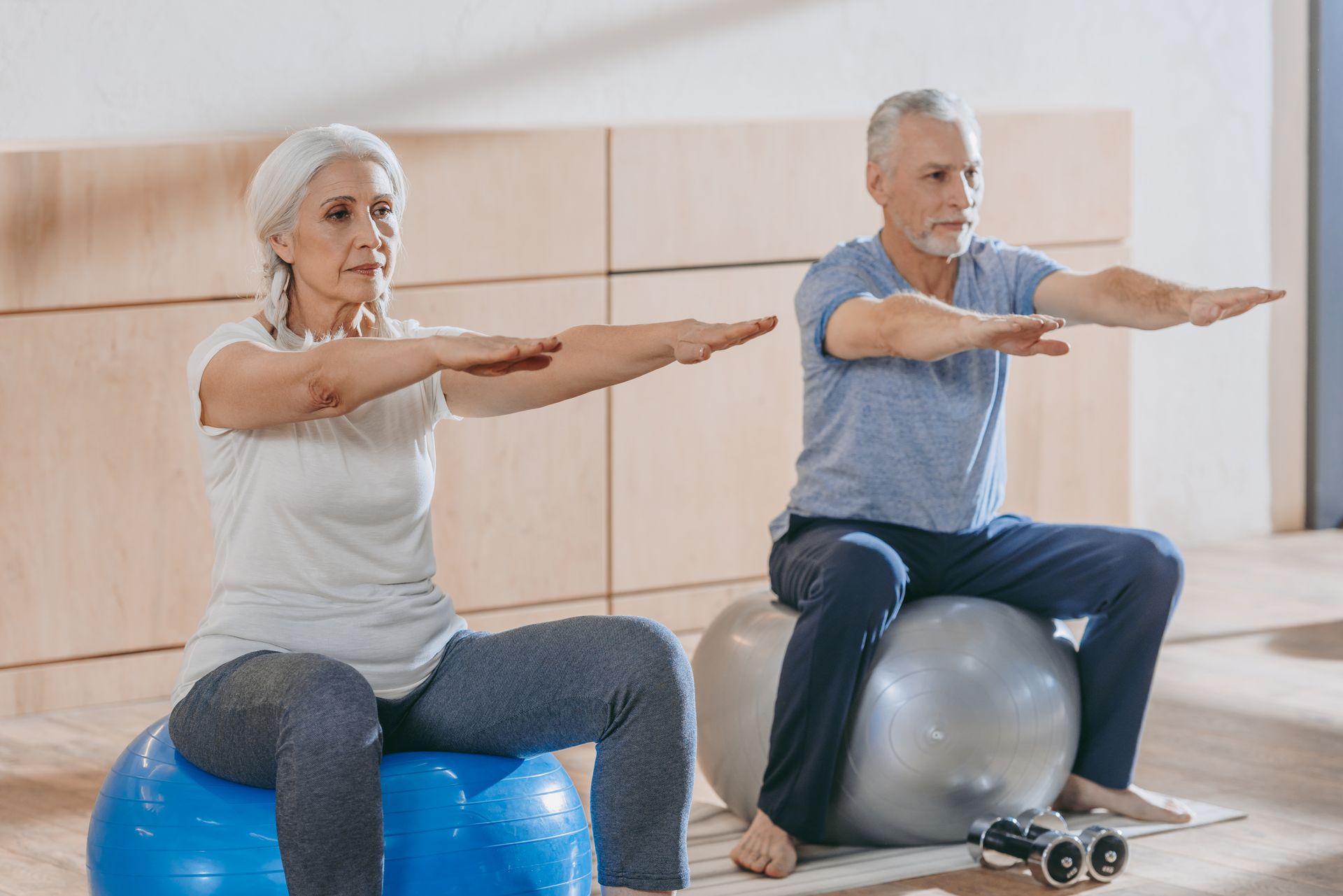 Woman and man on exercise balls, arms outstretched in a light-filled room.