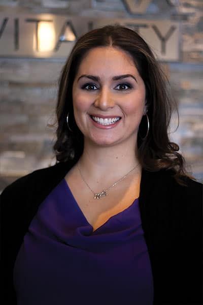 Woman with dark hair, smiling, wearing a purple shirt, black jacket, and hoop earrings, in front of a brick wall with the word